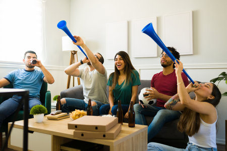 Cheerful sports fans using a vuvuzela and watching soccer on tv while drinking beer and eating pizzaの素材 [FY310187550046]