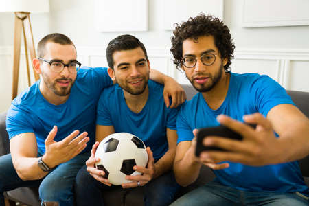 Handsome friends taking a selfie with a smartphone while enjoying watching a soccer game on tv