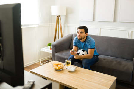 Handsome young man using the remote and watching the soccer game on TVの写真素材