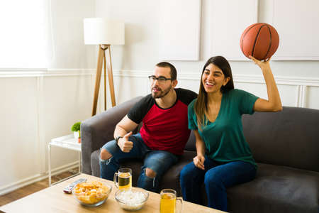 Happy young couple laughing while watching together the basketball game on the sofaの写真素材