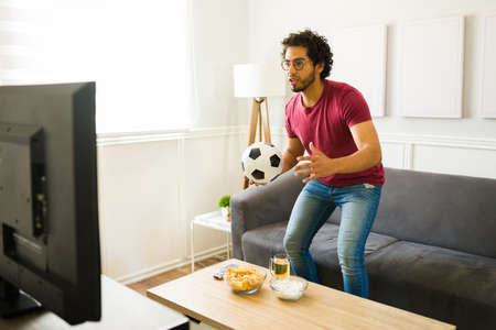 Worried young man standing up and feeling emotional in the living room while enjoying a soccer game on televisionの写真素材
