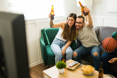 Attractive happy couple hugging and drinking beer while hanging out with friends in the living roomの写真素材