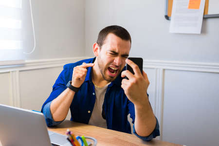 Angry young man calling his boss and screaming to his smartphone while working from homeの写真素材