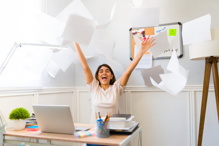 Excited young woman throwing papers and celebrating finishing her work while sitting at her desk at homeの写真素材