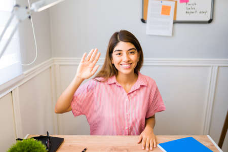 Attractive latin woman waving and saying hi while speaking with her boss during a work video call for her telecommute jobの写真素材