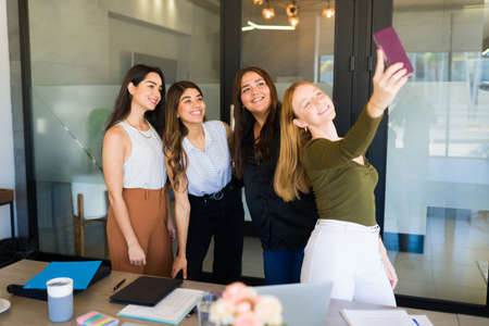 Attractive young woman feeling happy while taking a selfie with her female co-workers and friends at the officeの写真素材