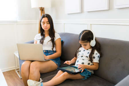 Busy young mom working from home and typing on the laptop with her young kid playing video games on the tabletの写真素材