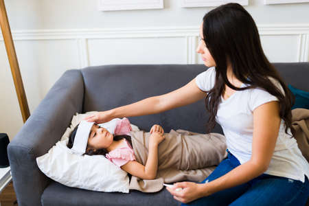 Sick young child with a fever and a cold resting on the sofa with her worried hispanic mom at home