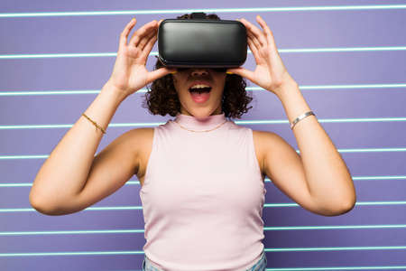 Surprised young woman looking excited while wearing VR glasses and using virtual reality technology against a futuristic backgroundの写真素材