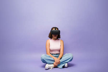 Brunette young woman with virtual reality glasses sitting alone on the floor and living through a metaverse simulationの写真素材