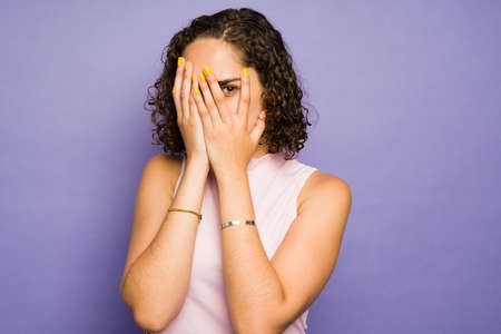 Shy woman with curly hair hiding her face with her hands and opening one eye while feeling afraid or...