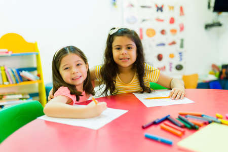 Multiracial girl friends smiling and hugging while sitting together at a desk in kindergarten schoolの写真素材