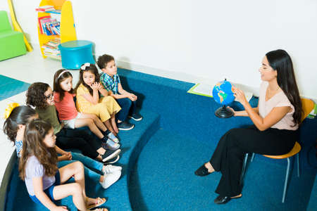 Attractive latin preschool teacher teaching a geography class and using a globe during a lesson with...