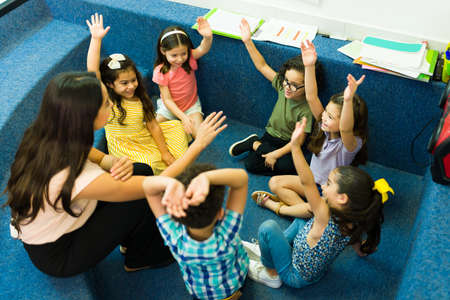 High angle of a female teacher giving a lesson to her kindergarten students raising their hands to...