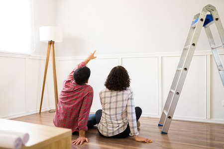 Young couple seen from behind sitting on the floor and dreaming about their new space while planning home renovationsの写真素材