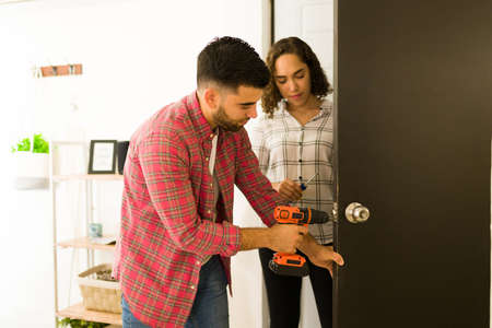 Handy young man and woman putting a new security doorknob with a drill on a doorの写真素材