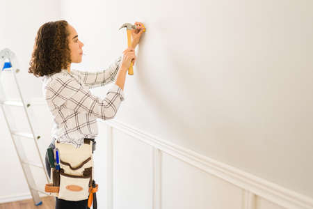 Young caucasian woman using a hammer and preparing to put a painting on the wall while doing home repairsの写真素材