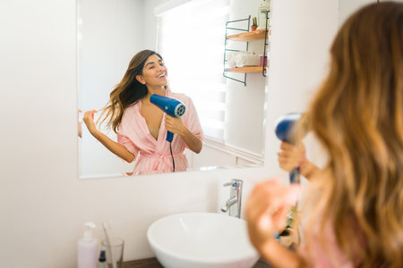 Gorgeous excited woman laughing while drying her hair with a blow dryer and getting ready in the bathroomの写真素材