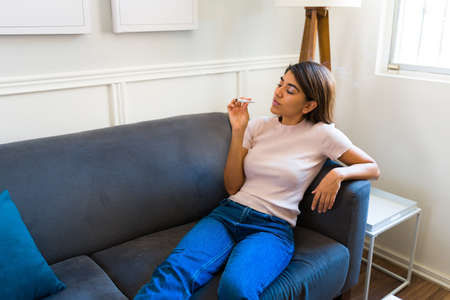 Latin young woman lying on the sofa at home while smoking cannabis to help with her pain or anxietyの写真素材