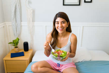 Excited attractive woman looking at the camera while eating a delicious green salad while sitting in...