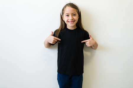 Portrait of a beautiful little girl looking happy pointing at the front design of her black mockup...