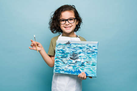Portrait of a smiling little boy holding his painting and paintbrush while learning to paint to become an artistの写真素材