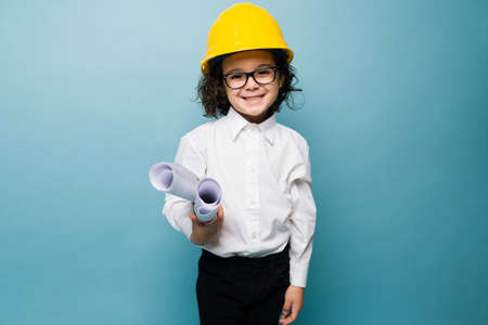 Happy little boy with glasses and a yellow helmet using construction plans while playing architect against a blue backgroundの写真素材