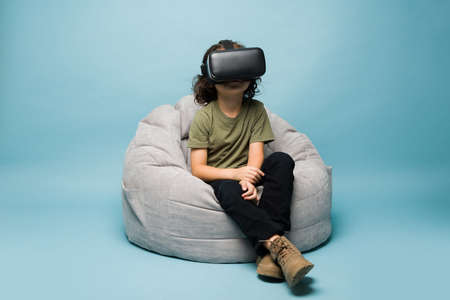 Caucasian elementary boy sitting on a kids chair and wearing virtual reality glasses and playing with futuristic technologyの写真素材