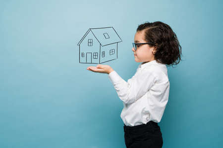 Cute caucasian boy with glasses playing architect with a house drawing against a blue studio backgroundの写真素材