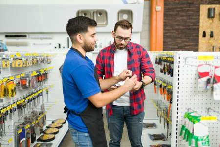 Happy young man working at the hardware store and helping a male customer looking to buy tools