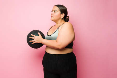 Overweight young woman working out with cross training and exercising using a slam ball against a...