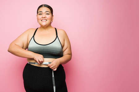 Upset obese woman feeling worried and stressed about her body weight while using a tape measure in front of a pink background with copy spaceの写真素材