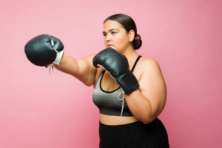 Overweight sporty young woman in activewear boxing using gloves and working out or training against a pink backgroundの写真素材