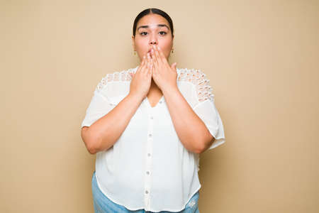 Studio shot of a surprised young woman covering her mouth and looking shocked against a yellow...