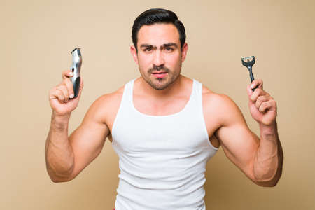 Worried young man in his 20s  grooming holding a disposable razor and an electric shaver while...