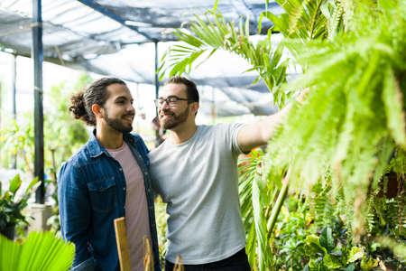 Handsome cheerful caucasian man hugging his gay partner while buying together plants at the nursery gardenの写真素材