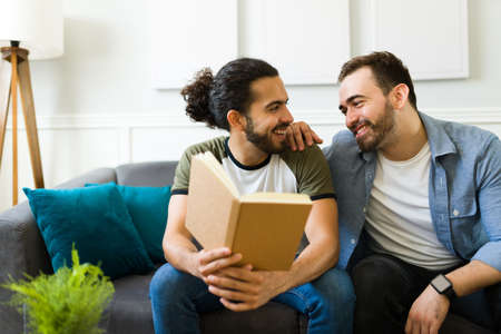 Cheerful gay man at home reading a book and laughing with his partner while having fun together during a relaxing timeの写真素材
