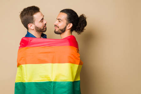 Romantic gay couple wrapped in a rainbow flag supporting the LGBT community rights while embracing against a background with copy spaceの写真素材