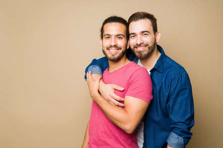 Attractive happy gay couple hugging and smiling while looking at the camera in front of a studio background with copy spaceの写真素材