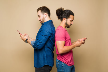 Profile of a caucasian gay man and hispanic boyfriend standing back to back texting on their smartphonesの写真素材