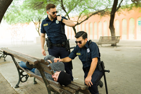 Concerned police officers trying to wake up a homeless man sleeping on a bench park to bring him to...