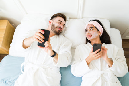 Cheerful smiling couple putting on a face mask and having a spa day together while relaxing in the bedroom using the smartphoneの写真素材