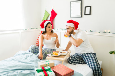 Attractive couple in pajamas wearing santa hats during christmas eating breakfast in bedの写真素材
