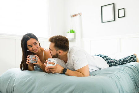 Beautiful happy woman and man looking at each other and smiling relaxing in pajamas while drinking coffee in bedの写真素材