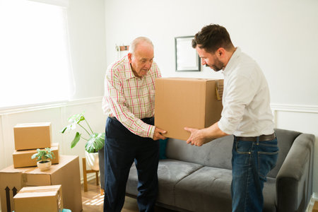 Young man carrying boxes and helping his elderly caucasian father to move to a new apartment