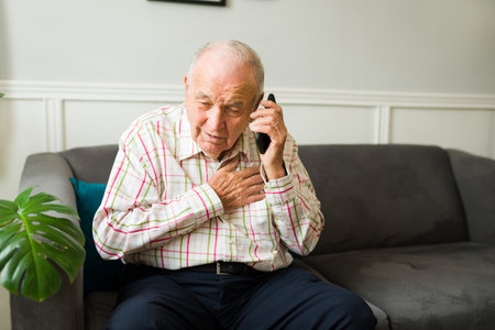Sad senior old man feeling very sick and having breathing problems while talking on the phone with...