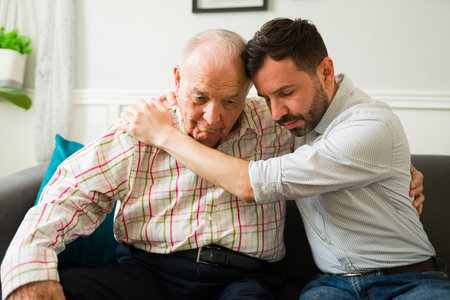 Sad young man missing his old senior father and hugging him while looking sad together at homeの写真素材