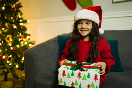 Cheerful young girl with a santa hat sitting on the sofa opening a christmas gift at nightの写真素材