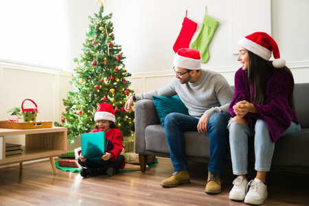 Cute cheerful little girl sitting under the christmas tree opening surprising presents with her parentsの写真素材