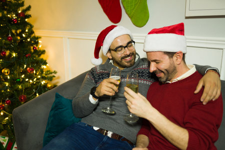 Excited gay couple laughing and having fun while drinking wine celebrating the christmas holidays at homeの写真素材